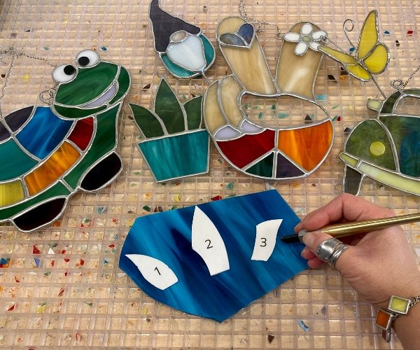 Variety of stained glass suncatchers including a gnome turtle peace sign plant and hand cutting pattern out of glass Mountain Woman Products Glass Classes