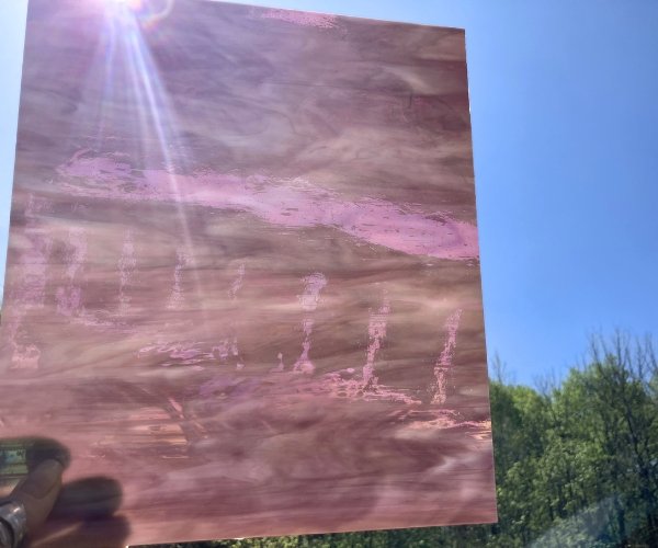 Designer Glass Iridized Amethyst stained glass sheet held up against a blue sky with trees a pinky type purple and white glass in the Mountain Woman Products studio DG1128