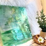 Designer Glass DG1075 Iridized Green & White stained glass sheet with a silvery color iridizing with a plant in front in Mountain Woman Products Studio