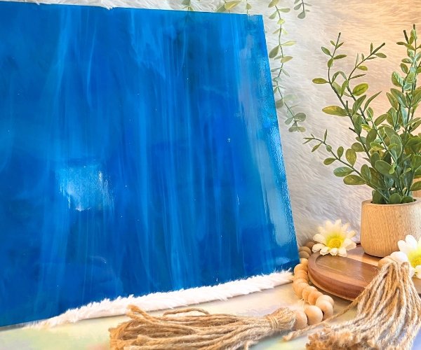 Oceanside Deep Aqua White Wispy Stained Glass Sheet is a darker blue color with white streaks with a plant and daisies in the foreground Mountain Woman Products