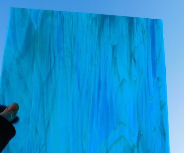 Oceanside Deep Aqua White Wispy Stained Glass Sheet is a darker blue color with white streaks being held by a hand in front of the sun Mountain Woman Products