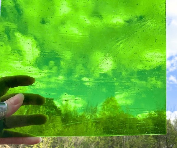 Designer Glass Medium Green Transparent Fusible COE 96 glass held up by hand with blue sky fluffy clouds and trees in background at the Mountain Woman Products studio
