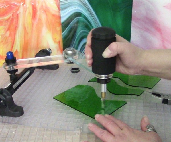 Creator's Cutter's Mate Mini Glass Cutter showing a hand using it to cut curves in green glass on a waffle grid with orange, teal, & pink glass in the background in the Mountain Woman Products studio
