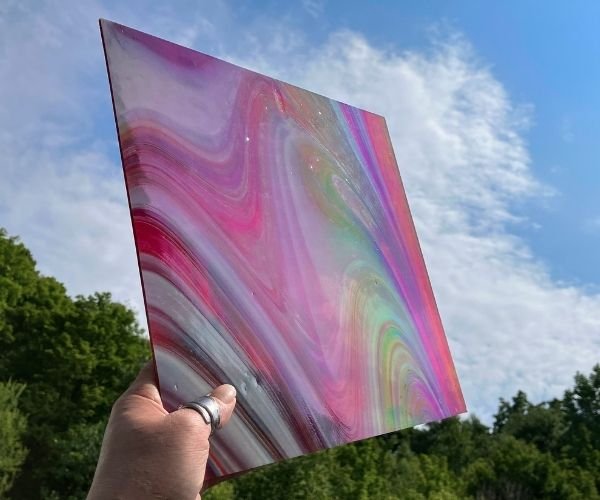 front of Oceanside Witching Hour Fuser's Reserve Special Edition Stained Glass Sheet showing red and white streaks with rainbow iridized on front against the backdrop of a blue sky and clouds at the mountain woman products studio