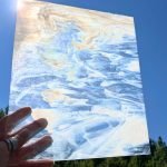 Designer Glass Clear & White Streaky stained glass sheet held in hand in front of blue sky with sun shining through at the Mountain Woman Products studio