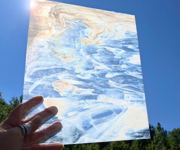 Designer Glass Clear & White Streaky stained glass sheet held in hand in front of blue sky with sun shining through at the Mountain Woman Products studio