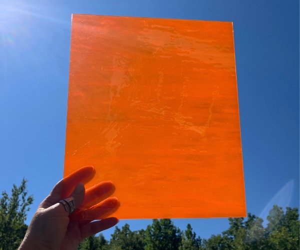 Designer Glass Dark Orange Transparent Fusible Glass with the sun shining through and sky and trees in background at the mountain woman products studio